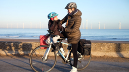 A pregnant woman stood smiling on a bicycle on the sea front in a helmet and coat while looking down at her toddler who is sat on the front of the bike and smiling up at her