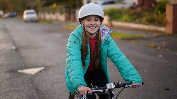Girl in blue coat and white helmet smiling at the camera while she cycles to school