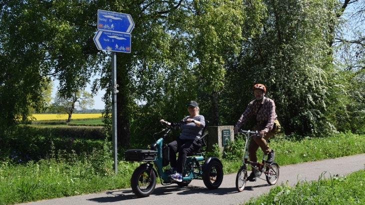 Two people traveling along a national cycle network path. The person on the left is using an electric trike and the person on the right is using a standard cycle. Around the path is lost of greenery and on the side of the path is a national cycle network sign.