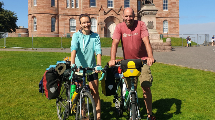 A man and a woman stood smiling with their bikes and paniers in the sunshine in front of a stately home in the UK 