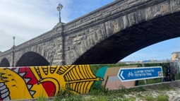 A colourful mural, displaying a yellow dragon beside a blue National Cycle Network sign for the Ystwyth Trail, is photographed with Pont Trefechan above it in the background.