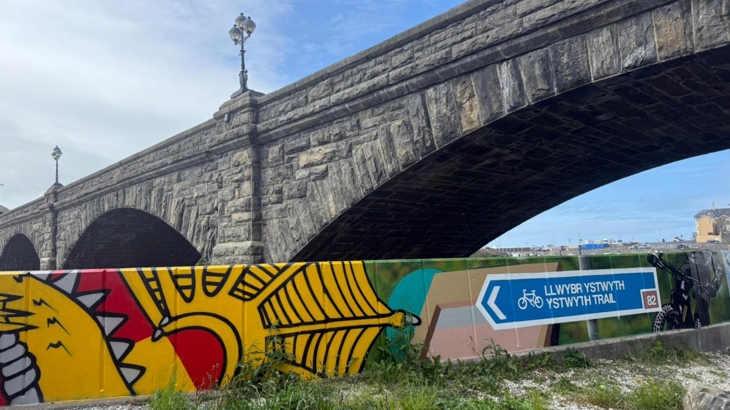 A colourful mural, displaying a yellow dragon beside a blue National Cycle Network sign for the Ystwyth Trail, is photographed with Pont Trefechan above it in the background.
