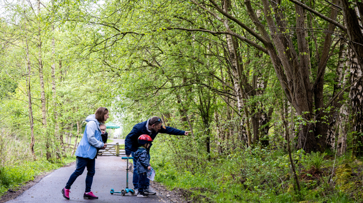A family of three with a young child on a scooter on a traffic-free path surrounded by greenery the man is pointing towards a hedge 