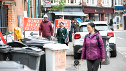 A sidewalk lined with bins, road signs and cars parked, with pedestrians navigating their way around them.
