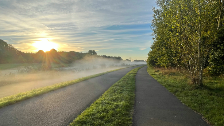 Cycle path along the River Exe, Exeter. The sun is rising and there is a low mist over the river and woodland.