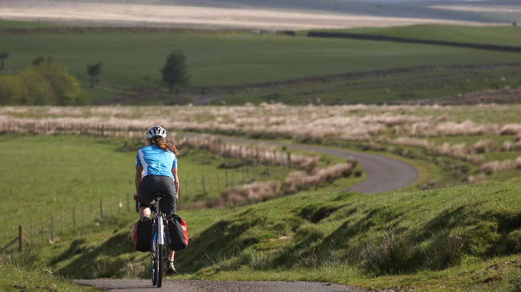 A woman on a cycle with two red panniers, with her back to the camera cycling down a winding hill on a road on Hadrian's Cycle Way
