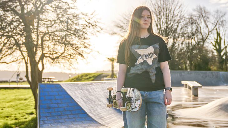 A teenager stood in front of a skate park holding a skateboard. Behind them are some trees.