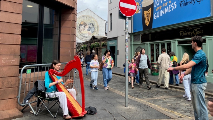Busker playing red harp on corner of Hill Street at Belfast music festival. Mural and street of people passing.