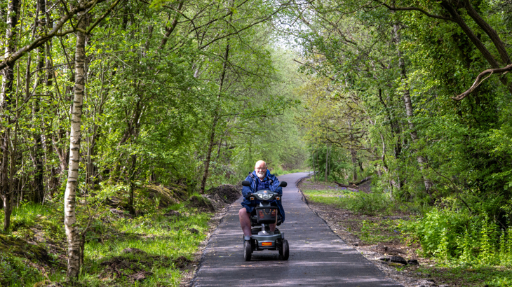 A man with grey hair in a mobility scooter wheeling along the National Cycle Network surrounded by greenery