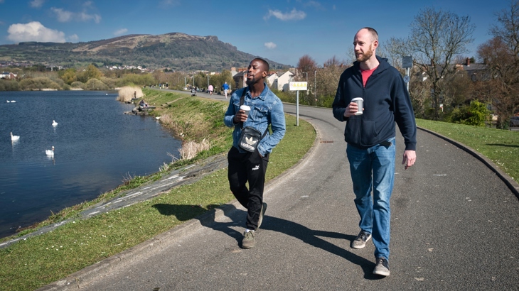 Two people walking with a coffee in hand along a like with a hill in the background.