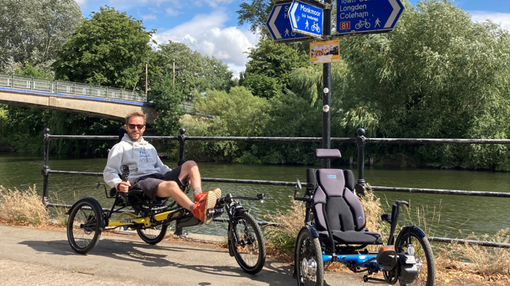 A man smiling in the sunshine with sunglasses on wearing shorts and sat on an adapted trike on the National Cycle Network next to a waterway