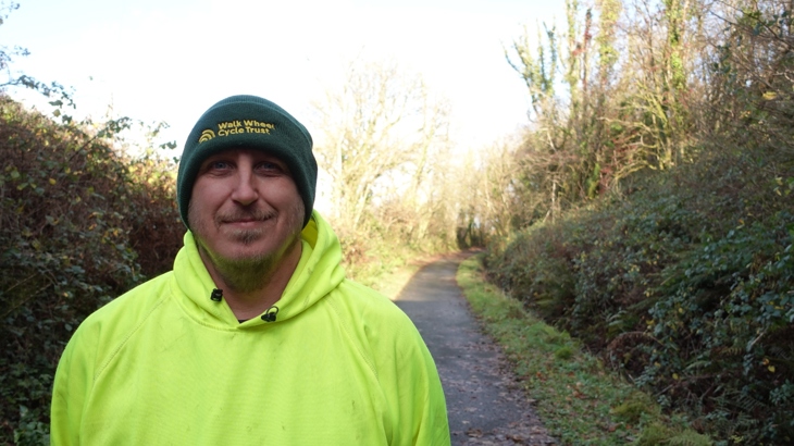 A man standing in front of a NCN path smiling and wearing a dark green "walk wheel cycle trust" beanie hat, and a bright green hoodie.
