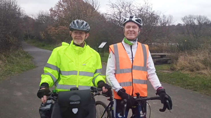 Two men smiling in high-vis and helmets stood on a traffic-free path with their bikes 