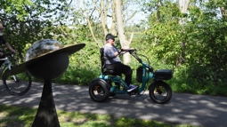 A person traveling along a national cycle network path on an electric trike. Around the path is lots of greenery, and in the foreground is a sculpture of a planet, signifying that the path is part of the "Solar System Way", another name for the York-Selby railpath.