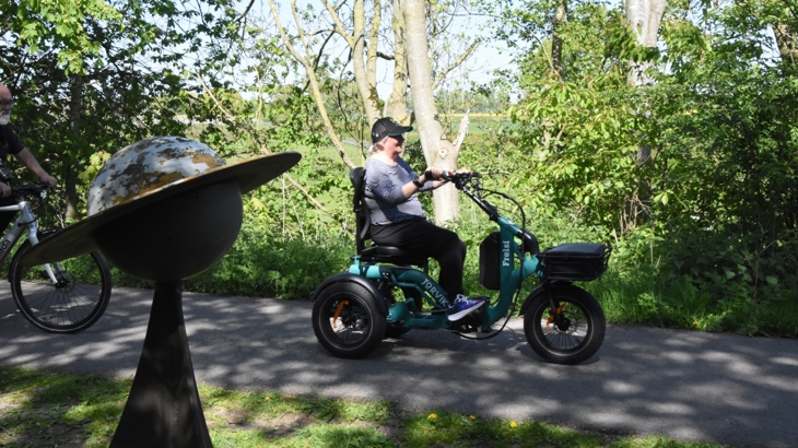 A person traveling along a national cycle network path on an electric trike. Around the path is lots of greenery, and in the foreground is a sculpture of a planet, signifying that the path is part of the "Solar System Way", another name for the York-Selby railpath.