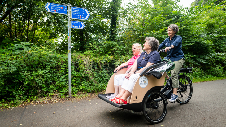 Two women sat in a trailer at the front of a bike being pedalled by a woman all smiling on a rural part of the Network