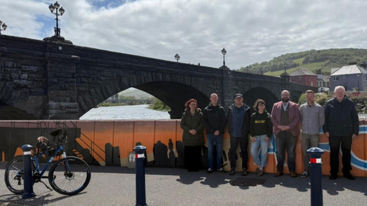 Walk Wheel Cycle Trust staff stand in front of an orange mural painted on a floodwall. It is sunny and cloudy, with Pont Trefechan pictured in the background.