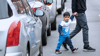 Little boy crosses a city road, lined with parked cars, holding his dad's hand and looking earnestly at the camera