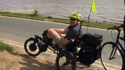 A man sat on a recumbent trike on the side of a road smiling with sunglasses on. There's a body of water on the other side of the road