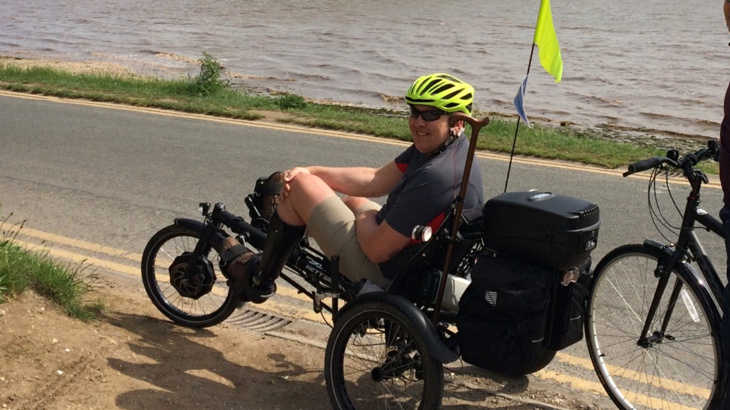 A man sat on a recumbent trike on the side of a road smiling with sunglasses on. There's a body of water on the other side of the road