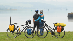 Two people standing together with their bikes, facing towards a body of water.