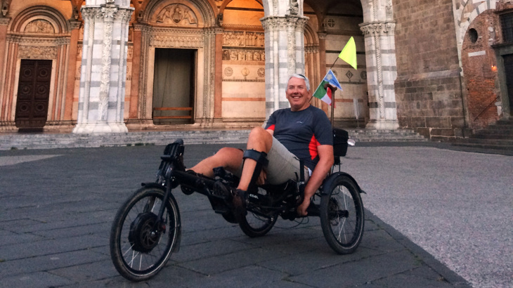 A man with short white hair wearing a t-shirt and shorts sat on a recumbent trike smiling in a European city 