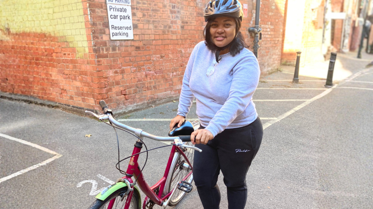 A person wearing helmet and exercising clothes stands in front of a building with their new red adult bike. 