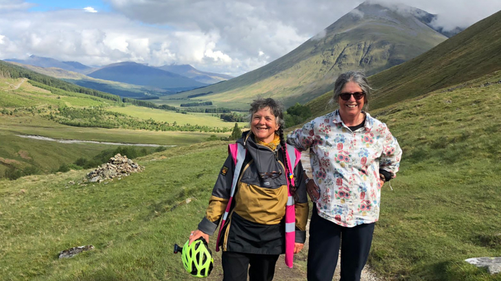 Two women stood smiling side by side against the rolling green hills of Scotland on a bright day