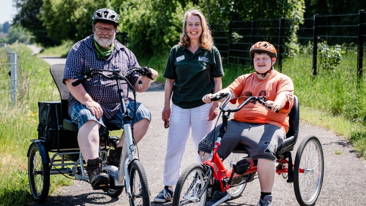 Two cyclist on tricycle and a person standing in between them. All smiling at the camera.