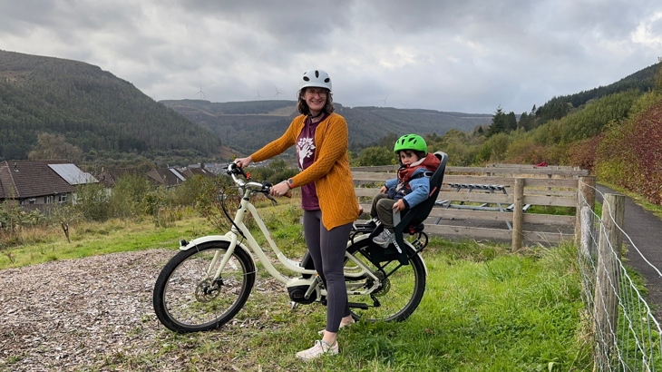 A mother stands astride her e-bike with her young son sat on the back, with a view of a town in a valley in the background.