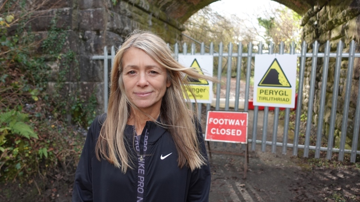 A person with long blonde hair smiling in front of a path that has been close off. Behind them is a sign that reads "footway closed".