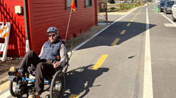 A man sat in an adapted cycle in the sunshine wearing a helmet on a long stretch of tarmac