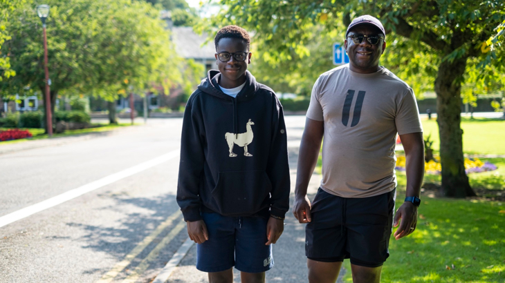 A father and son stood side by side smiling on a sunny day surrounded by green trees 