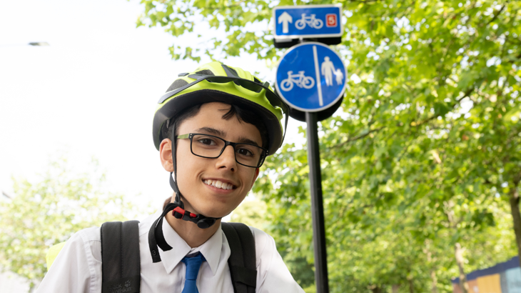 James smiles at the camera, wearing a yellow cycle helmet and a rucksack over a school shirt. Bright green trees and a blue cycling walking sign are in the background.