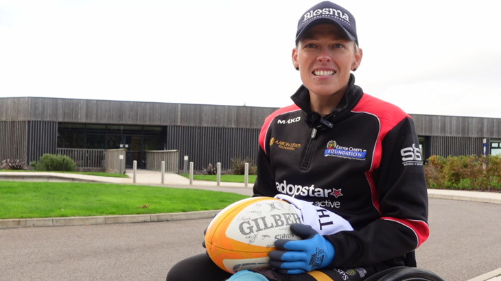 A woman in a wheelchair smiling while wearing a cap and holding a rugby ball in her lap