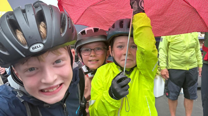 Three young boys wearing helmets and raincoats stood underneath an umbrella on a rainy day in Scotland 