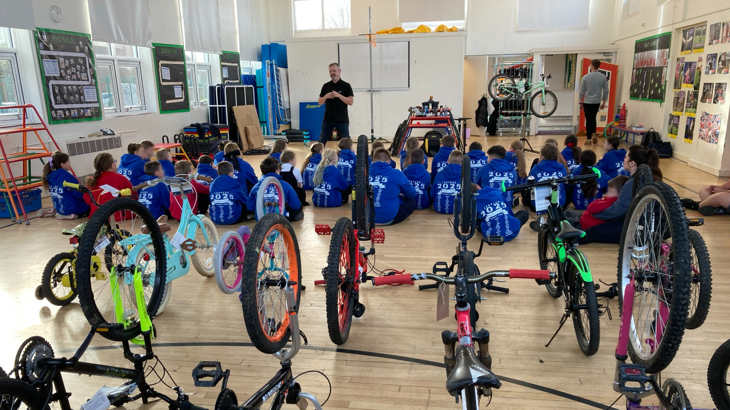 A school hall filled with primary school children with their backs to the camera and small bikes upside down with their wheels in the air while a man talks to the seated children at the front of the hall