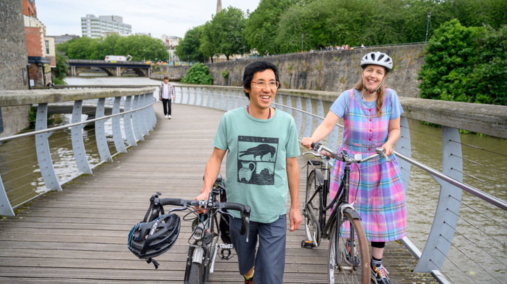 Two people in bright clothing, smiling and walking alongside their bikes on a bridge in Bristol city centre on a cloudy but bright day
