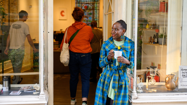 Person wearing a blue and yellow tartan coat standing in the doorframe of a cafe holding a cup, smiling. 